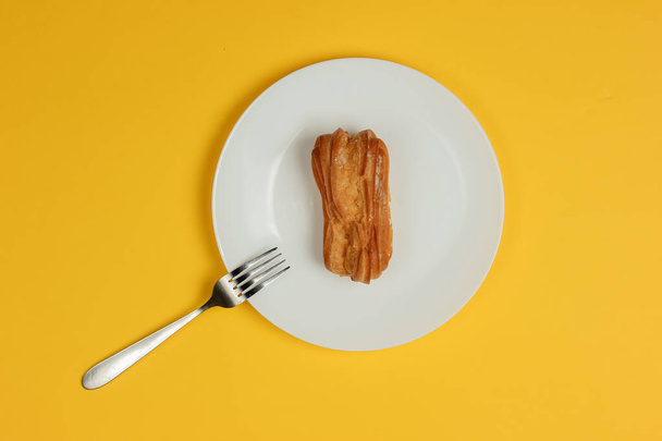 Eclair in plate with fork on yellow background. Top view - 写真・画像