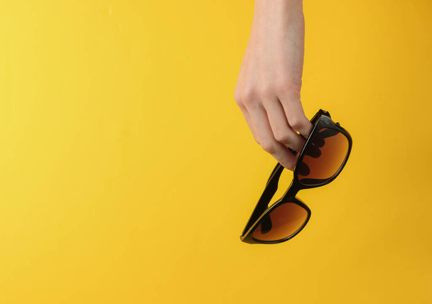 Female hand holds stylish sunglasses on a yellow background. Fashion studio shot - Foto, Imagen