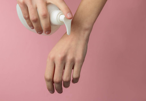 Woman holds cream bottle and applying moisturizer to beautiful hands for clean and soft skin. The concept of health and beauty. Pink pastel background - 写真・画像