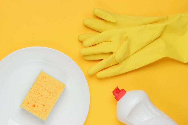 Set of tools for washing dishes on yellow studio background. Plate, rubber gloves, sponge, bottle. Top view - Photo, Image