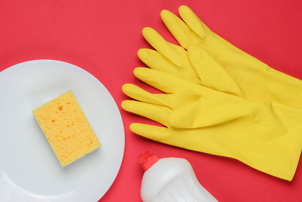 Gereedschap voor het afwassen van afwas op een rode studioachtergrond. Bord, rubberen handschoenen, spons, fles. Bovenaanzicht. Vlakke plaat - Foto, afbeelding