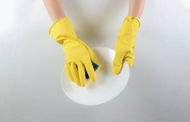 El concepto de una ama de casa lavar los platos. Las manos en guantes de látex amarillo lavan la placa con una esponja sobre un fondo gris - Foto, Imagen
