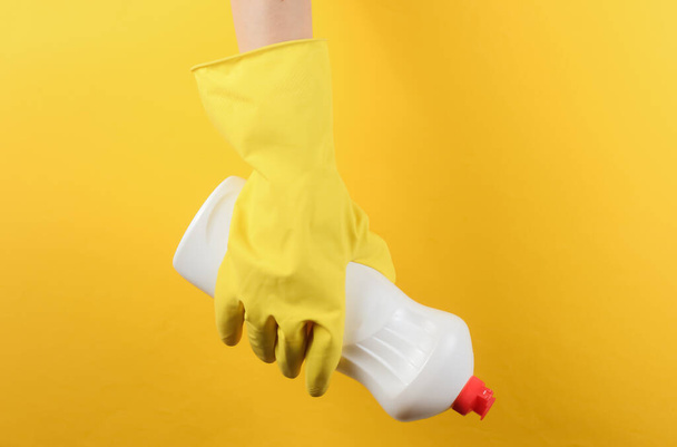 Női kéz sárga gumikesztyűben egy üveg mosószert tart sárga stúdió háttérben. Takarítási koncepció - Fotó, kép