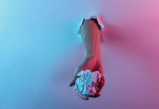 Hand holding crumpled paper ball through torn hole. Creative pop art pink blue neon color. Trendy gradient illumination. Night light - Foto, Imagen