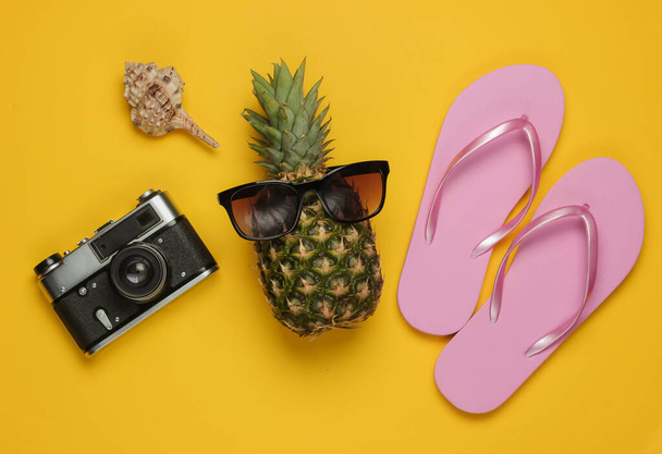 Fundo de verão. Diversão e humor. O conceito de umas férias de praia, viagem. Abacaxi com óculos de sol, chinelos, câmera no fundo amarelo. Vista superior - Foto, Imagem