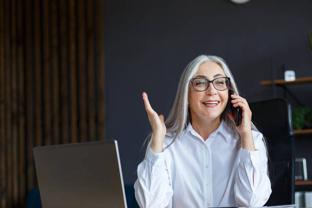 Smiling senior grey-haired beautiful businesswoman talking on the phone in office. Business portrait of attractive mature lady wearing eyeglasses sitting at workplace. Conversation, negotiation - Foto, imagen