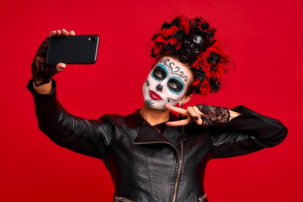 Dziewczyna z cukrem makijaż czaszki z wieńcem kwiatów na głowie i czaszce, nosi koronkowe rękawiczki i skórzaną kurtkę, dzięki czemu selfi izolowane na czerwonym tle. koncepcja Halloween lub La Calavera Catrina. - Zdjęcie, obraz