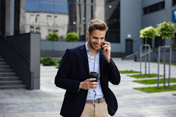 Portrait of handsome smiling man in casual wear holding smartphone. Successful manager makes a call. Young businessman talking on the phone, standing on city street near business center - Фото, изображение