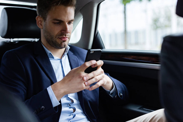 Close up portrait of young man in suit holding smartphone while sitting on the back seat of the car. Handsome young businessman test drive new car - Foto, imagen