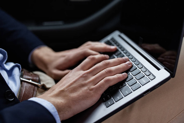 Close-up photo of male hands with laptop. Businessman in suit sitting in car, texting message, searching web, browsing information using laptop keyboard. Online education, remote working - Photo, Image
