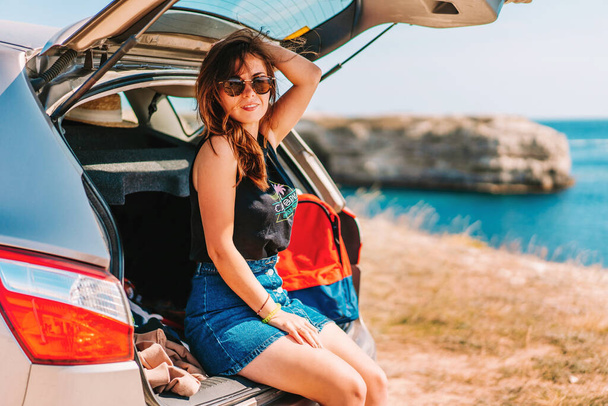 Молодая женщина в солнечных очках сидит на багажнике автомобиля и выглядит круто, на заднем плане море - Фото, изображение