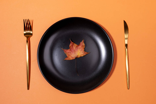 Autumn creative concept. Top view one dried colorful maple leaf on black plate with golden fork and knife on trendy pastel orange background. Autumn, fall, halloween, thanksgiving day concept. - Fotografie, Obrázek
