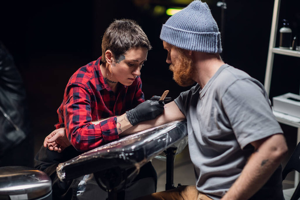 A young hipster man in a knitted hat makes a bull tattoo on his arm in a tattoo parlor with the help of a young girl master. - Фото, зображення