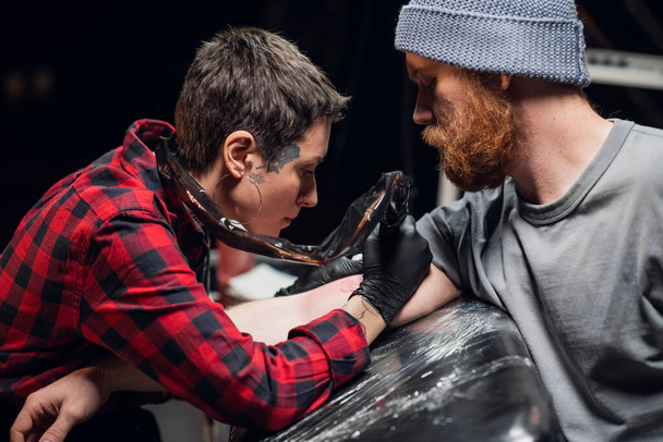 Dans un salon de tatouage moderne, une fille avec des piercings et des tatouages sur son corps fait un tatouage sur le biceps d'une jeune main hipster. - Photo, image