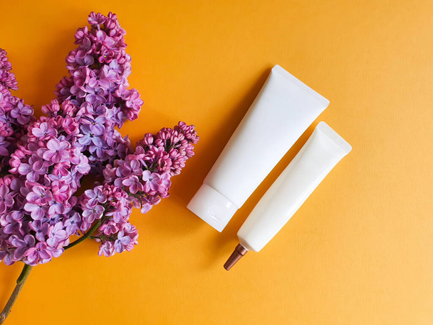 Макет двух небрендовых белых сжимающих косметических трубок для брендинга и лиловых цветов на ярко-желтом оранжевом фоне. Натуральная органическая косметика. Копирование пространства - Фото, изображение