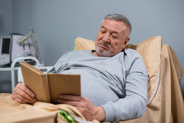 Ein grauhaariger alter Mann liest in seinem Bett auf einer Krankenstation ein Buch - Foto, Bild