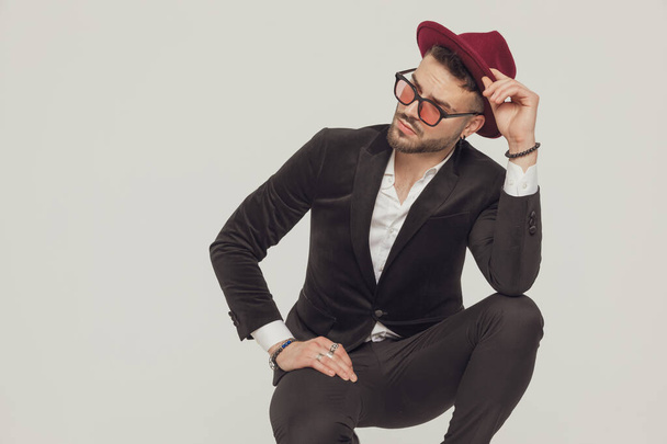 Pensive fashion model fixing his hat and looking away, wearing sunglasses while crouching on gray studio background - Foto, imagen