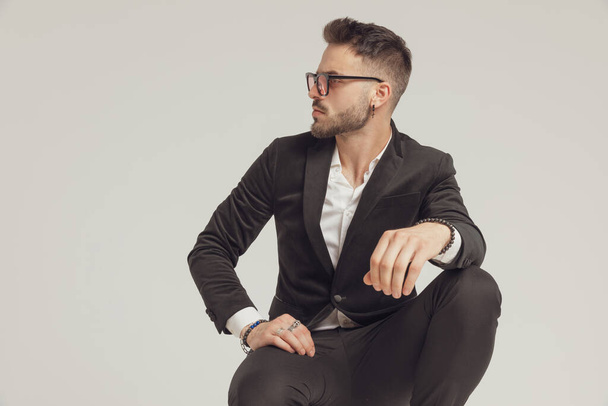 Confident fashion model curiously looking away and wearing sunglasses while crouching on gray studio background - Φωτογραφία, εικόνα
