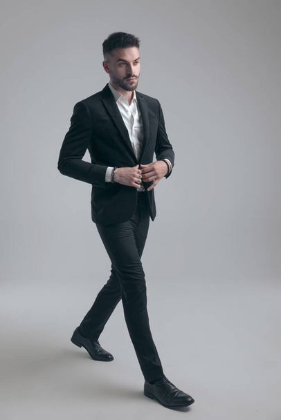 Charmant eleganter Mann knöpft seine Jacke auf, trägt Anzug, während er auf grauem Studiohintergrund läuft - Foto, Bild
