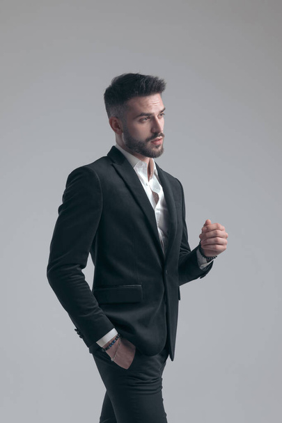 Eifriger eleganter Mann hält Hand in Tasche, trägt Anzug beim Gehen auf grauem Studiohintergrund - Foto, Bild