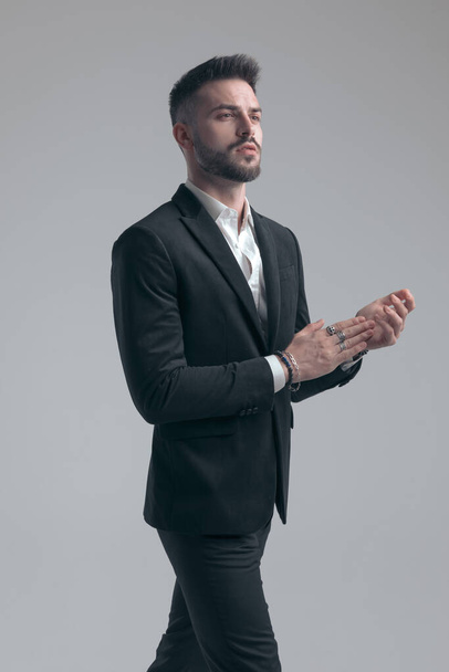 Stolzer eleganter Mann klatscht und schaut weg, trägt Anzug beim Gehen auf grauem Studiohintergrund - Foto, Bild