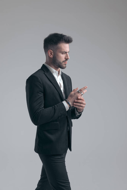 Hombre elegante confiado ajustando sus anillos, usando traje y caminando sobre fondo gris del estudio - Foto, imagen