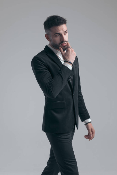 Eager elegant man holding hand on chin and looking away, wearing suit and walking on gray studio background - Foto, imagen