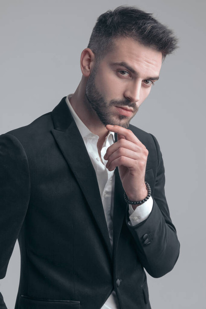 Γοητευτικός κομψός άντρας κρατώντας το χέρι στο πηγούνι και τη σκέψη, φορώντας κοστούμι ενώ στέκεται σε γκρι φόντο στούντιο - Φωτογραφία, εικόνα
