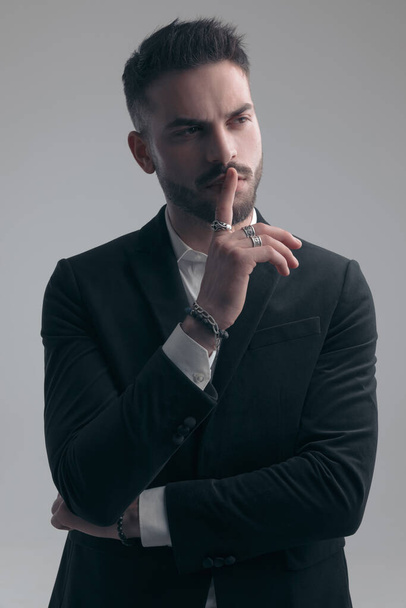 Mysterieuze elegante man gebaren heel, het dragen van pak terwijl staan op grijze studio achtergrond - Foto, afbeelding