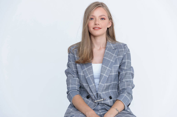 Positive businesswoman wearing suit and smiling while sitting on white studio background - Φωτογραφία, εικόνα