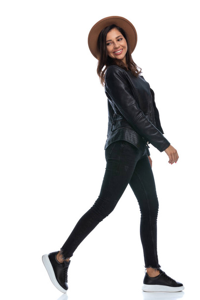 Side view of positive casual woman smiling and looking over shoulder, wearing hat and leather jacket while walking on white studio background - Photo, image