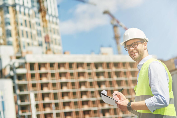 Молодой инженер-строитель или надзиратель в шлеме улыбается в камеру во время осмотра строительной площадки и делает заметки - Фото, изображение