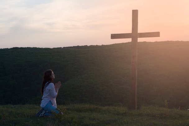 Молодая женщина на коленях. Молится. Руки в молитве. Возле деревянного креста. - Фото, изображение