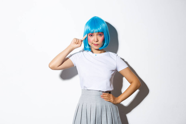 Imagen de chica asiática en peluca azul mirando escéptico, señalando con el dedo a la cabeza, de pie sobre fondo blanco - Foto, Imagen