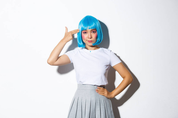 Изображение обеспокоенной азиатской женщины, выглядящей незабавно, направляющей ручной пистолет на голову, носящей синий парик на вечеринку в честь Хэллоуина, стоящей на белом фоне - Фото, изображение