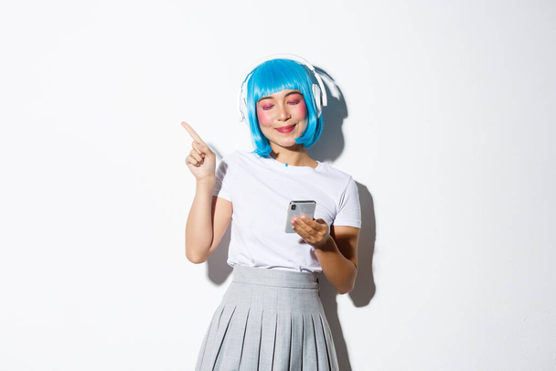Image de belle fille asiatique en perruque anime bleue, écoutant de la musique avec un sourire heureux, pointant du doigt le coin supérieur gauche et tenant smartphone - Photo, image