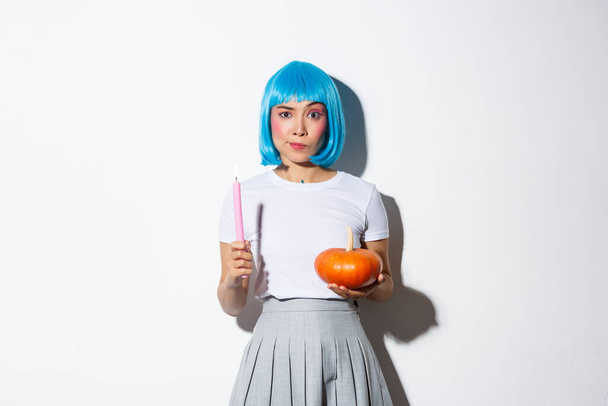 Konzept von Halloween. Bild eines attraktiven skeptischen asiatischen Mädchens mit blauer Perücke, das Kerze und Kürbis hält und ernst in die Kamera schaut - Foto, Bild