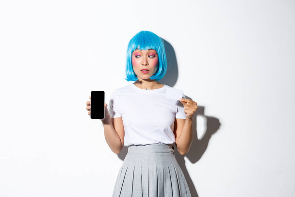 Epämääräinen kaunis aasialainen tyttö sinisessä peruukissa, katsot epävarma matkapuhelimeen, näyttää jotain näytöllä ja luottokortilla, seisoo valkoisen taustan yläpuolella - Valokuva, kuva