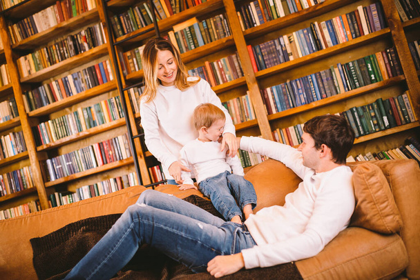 Christmas family portrait. Young Caucasian family in identical clothes having fun on sofa in living room with large library of books and Christmas tree in evening. Happy New Year and Merry Christmas. - Photo, Image