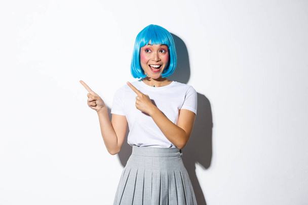 Opgewonden aantrekkelijke aziatische vrouw in blauwe pruik en schooluniform, wijzende vingers op de linker bovenhoek, op zoek geamuseerd en glimlachend over witte achtergrond - Foto, afbeelding
