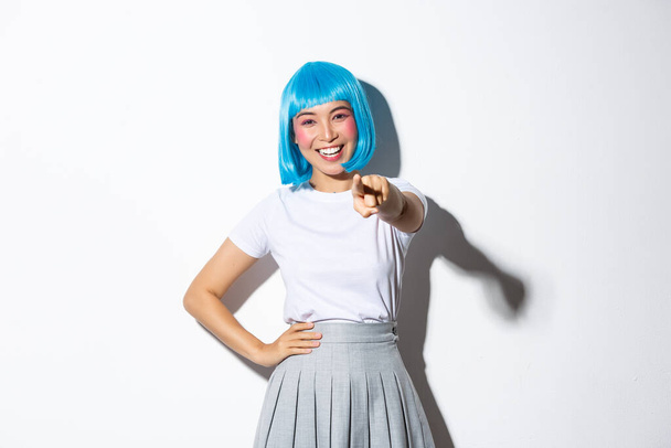 Portret szczęśliwej pięknej azjatyckiej dziewczyny uśmiechniętej i wskazującej palcem na kamerę, dokonującej wyboru, stojącej w niebieskiej peruce na białym tle - Zdjęcie, obraz