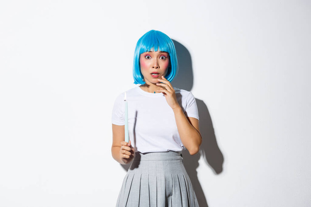 Concept d'Halloween. Image de fille asiatique timide effrayée en perruque bleue, tenant une bougie et regardant quelque chose d'effrayant - Photo, image