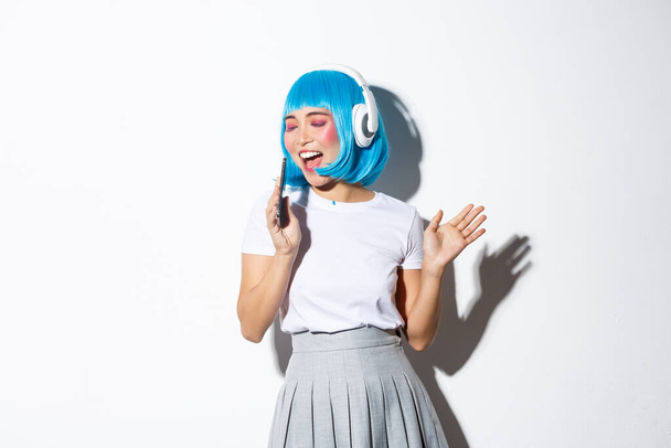 Imagen de hermosa chica asiática en peluca azul sintetizándose en la dinámica del teléfono móvil, usando auriculares, jugando aplicación de karaoke, de pie sobre fondo blanco - Foto, Imagen