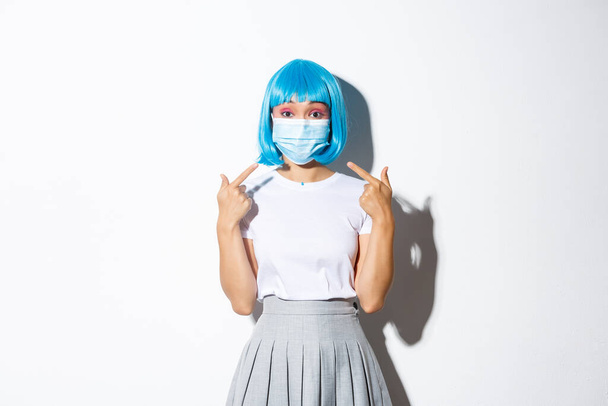 Concept of halloween celebration and covid-19 pandemic. Image of asian girl in blue wig pointing at medical mask, standing over white background - Photo, Image