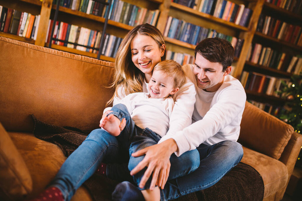 Happy parents playing with small child at Christmas time at home in living room on sofa near large cabinet with books and Christmas tree in evening. Family holiday in winter new year and christmas. - Photo, Image