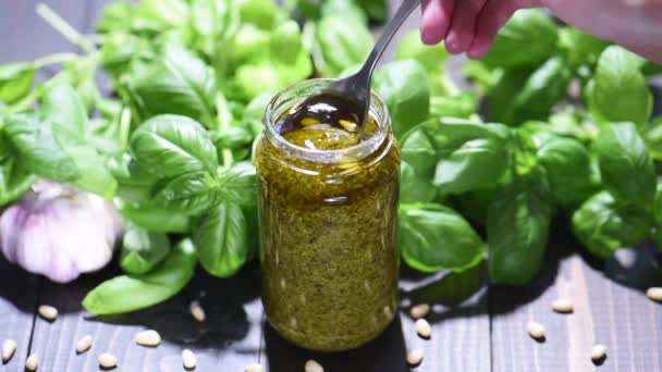 Small jar of homemade pesto sauce with green fresh sweet basil on wooden table. Woman's hands holding spoon - Footage, Video