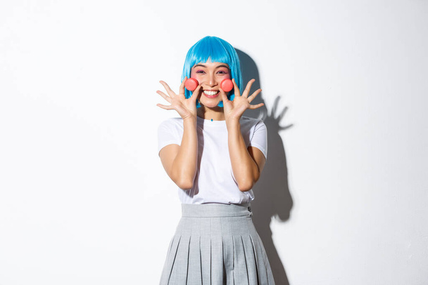Afbeelding van mooie glimlachende aziatische meisje kijken dom, tonen macarons, het dragen van blauwe anime pruik, staande over witte achtergrond - Foto, afbeelding