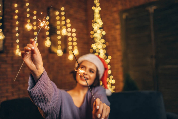 Pretty happy young brunette in christmas hat with sparkler in hands - Photo, image