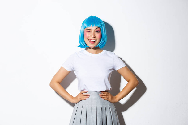 Portrait de belle fille asiatique en perruque de fête bleue montrant la langue et cligner des yeux stupide, debout sur fond blanc en costume d'Halloween kawaii - Photo, image