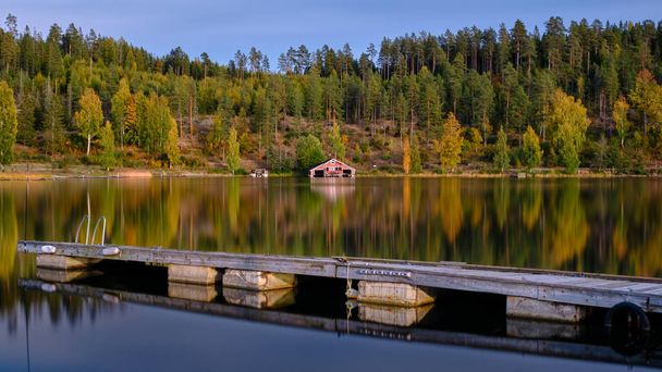 Jetty in the foreground and a red boathouse and forest in autumn colors in the background - Фото, изображение